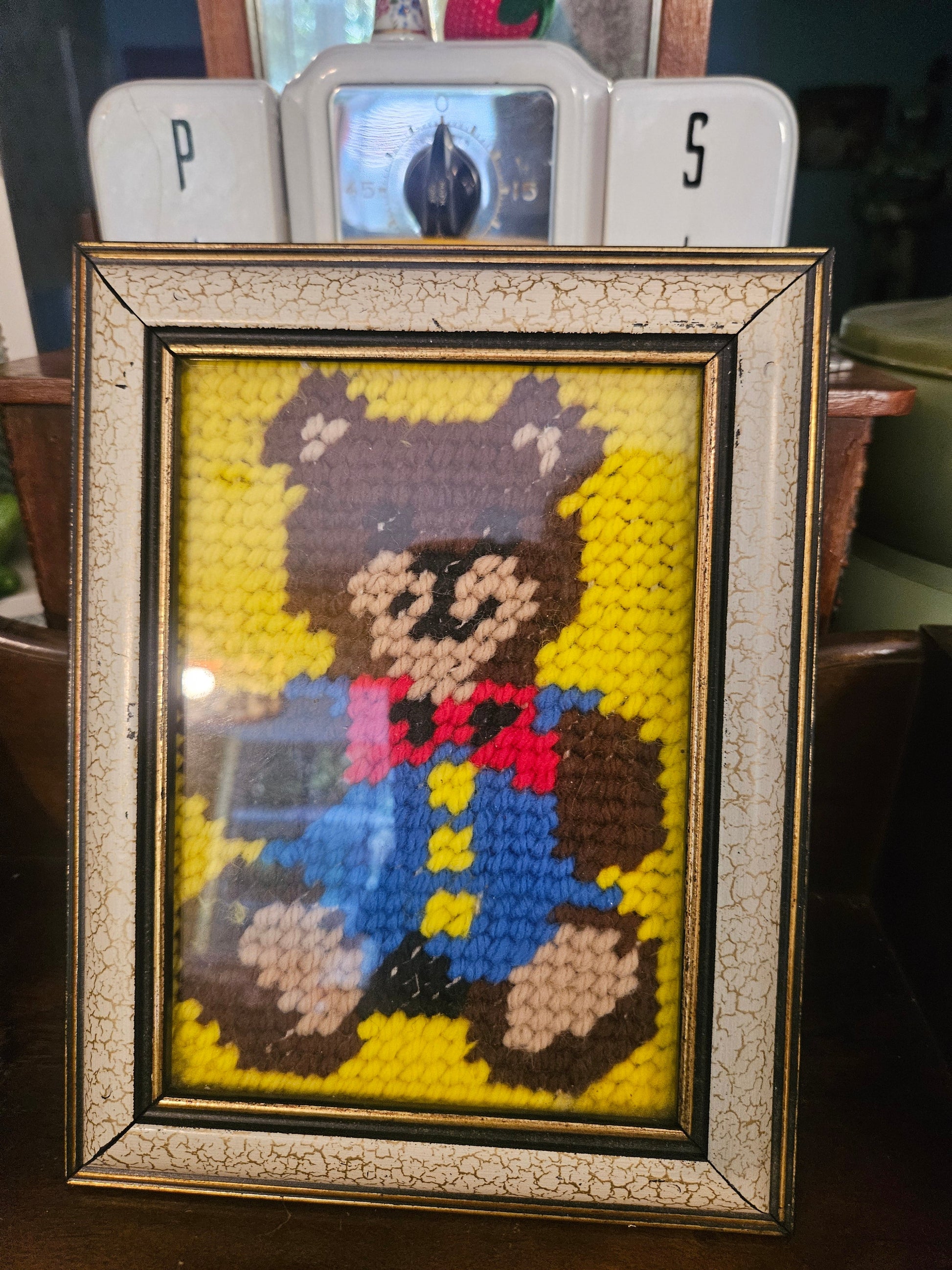 Vintage framed needlepoint teddy bear in blue overalls and red bow tie on yellow background in crackle gold frame