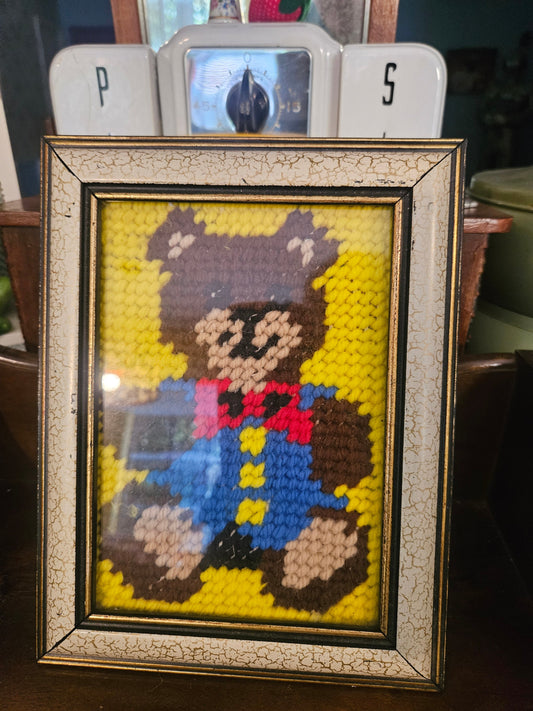 Vintage framed needlepoint teddy bear in blue overalls and red bow tie on yellow background in crackle gold frame