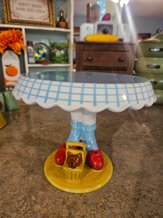 Wizard of Oz Dorothy legs ceramic cake stand front view showing gingham scalloped rim ruby slippers and Toto in basket
