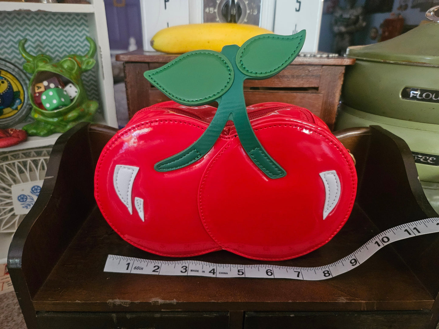 Red patent double-cherry novelty crossbody bag front view showing green leaf stem appliqué and white highlights with ruler for scale