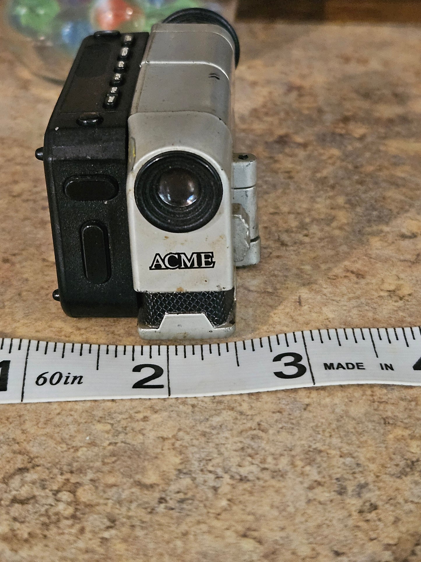 Front of ACME miniature camcorder showing lens and ACME logo with ruler for scale