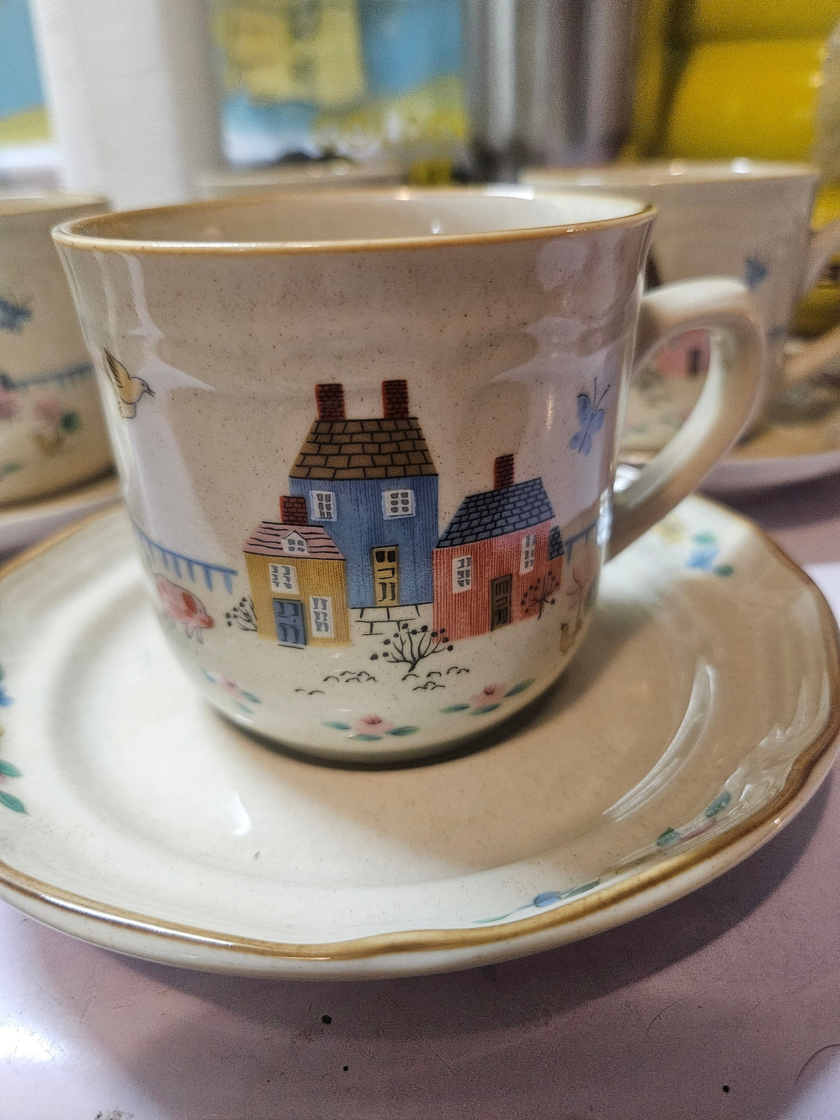 Vintage Heartland stoneware cup with folk-art village scene in pastel houses, birds, and butterflies, resting on a gold-trimmed saucer with floral accents