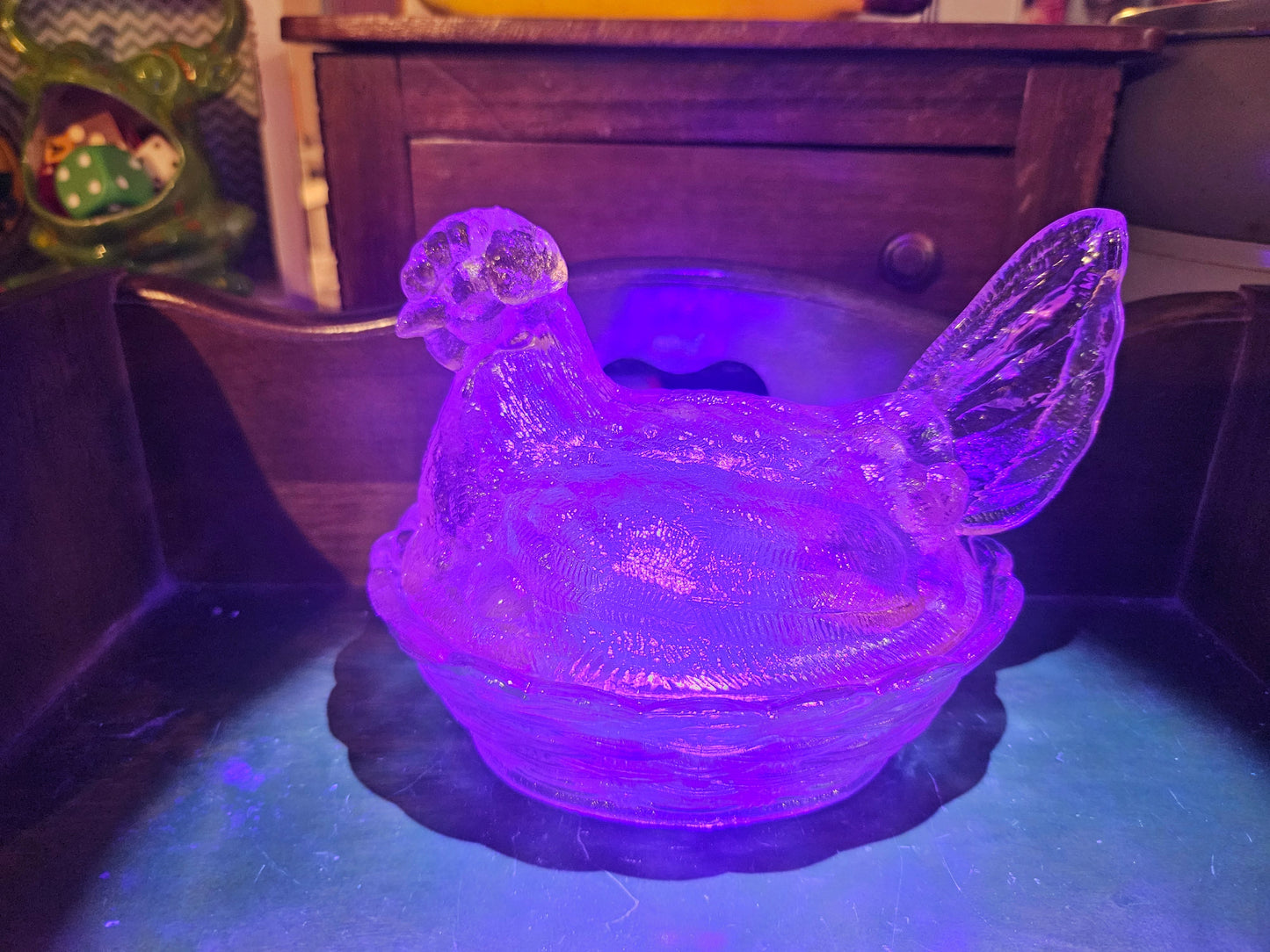 Clear pressed glass hen on nest glowing vivid purple under UV blacklight indicating uranium or manganese glass