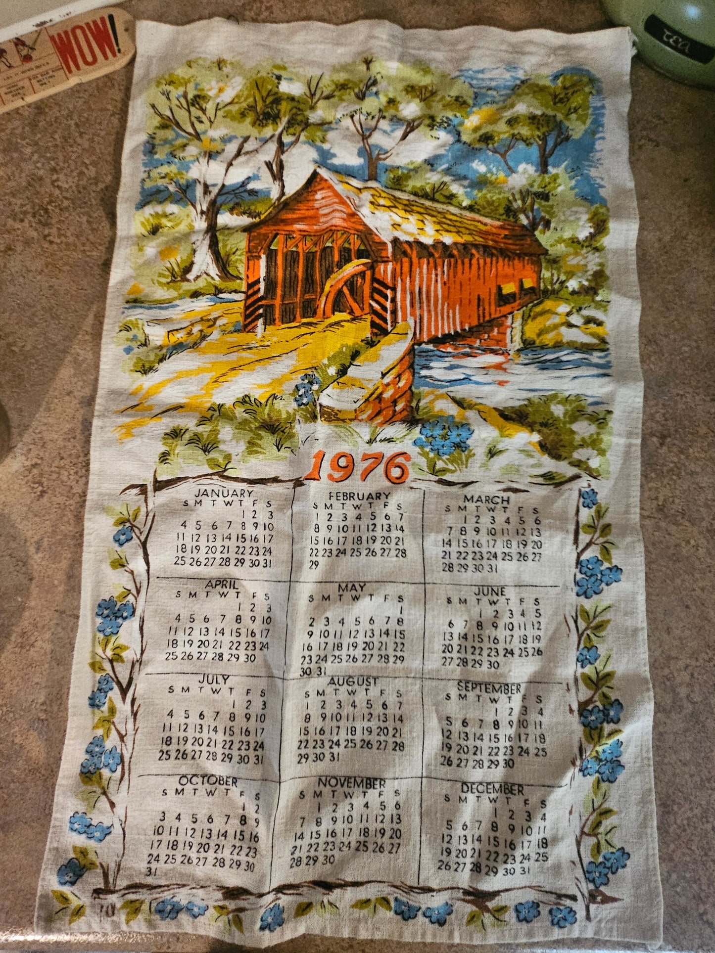 1976 covered bridge calendar tea towel showing full-color scene with waterwheel, forget-me-nots, and 12-month calendar